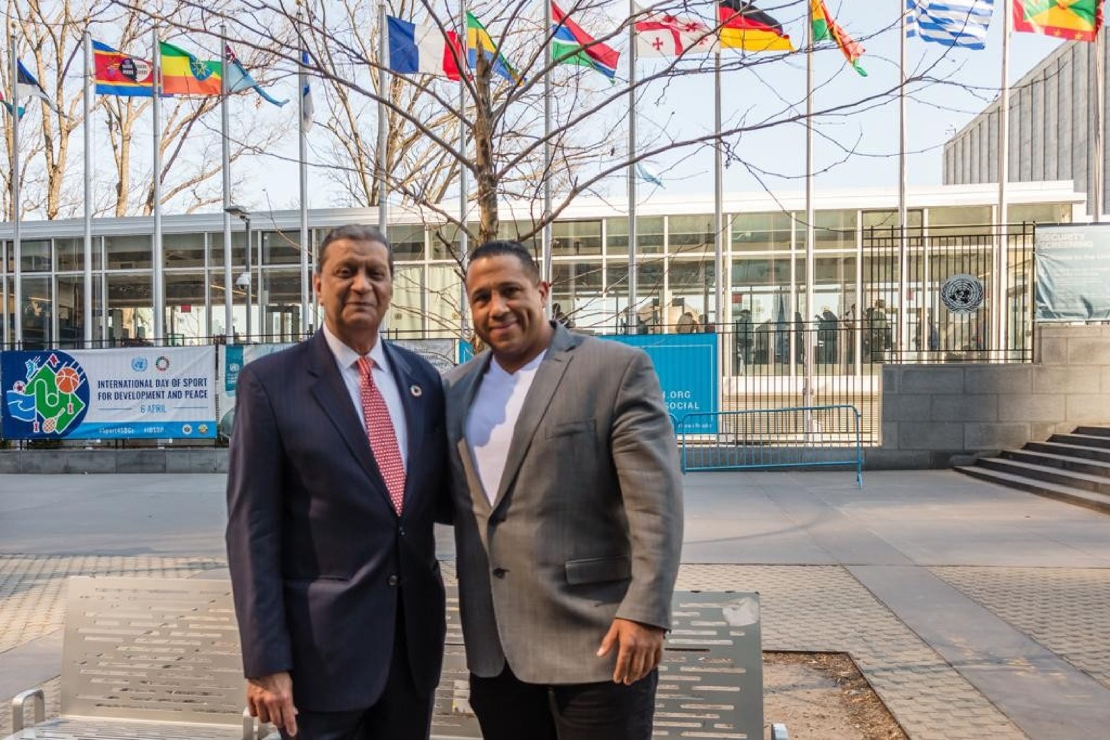 Asa Saint Clair and Amir Dossal standing together outside UN headquarters in New York, flags of member states and the banner for the International Day of Sport for Development and Peace behind them.