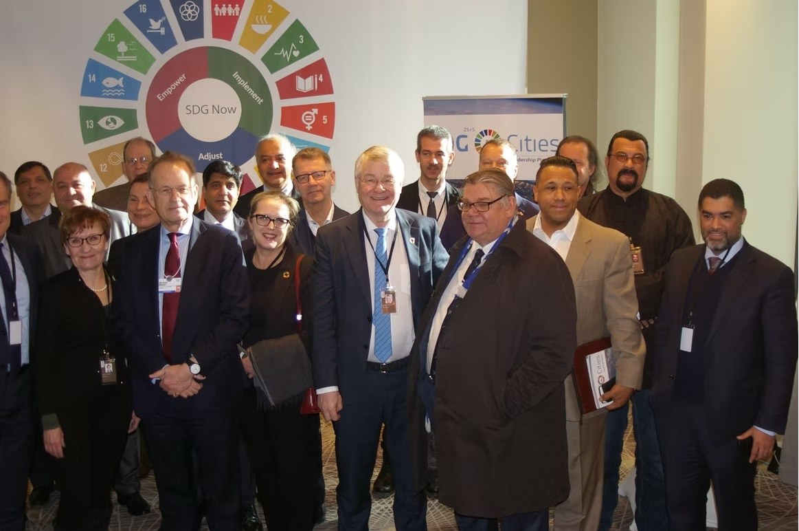 Group photo at an SDG Cities event beneath the SDG Now wheel. Michael Møller stands in the front row, left of center, under the word Empower; Asa Saint Clair stands at right, in a tan jacket.