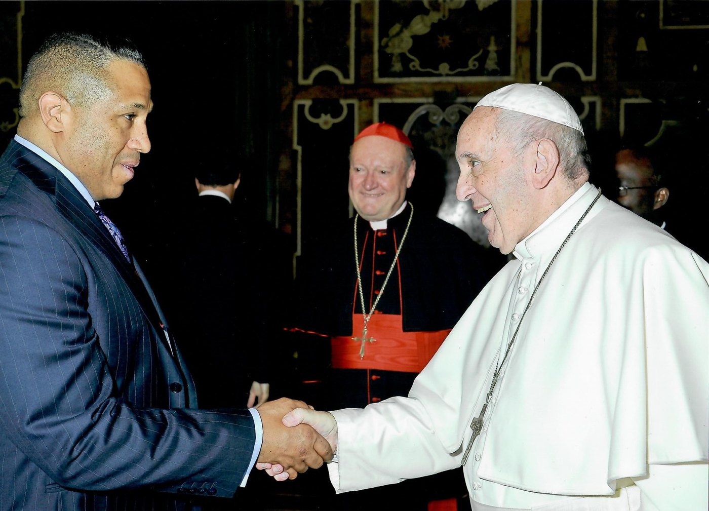 Asa Saint Clair shaking hands with Pope Francis at the Vatican, a cardinal smiling behind them.