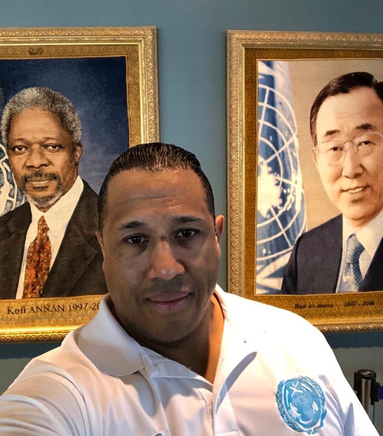 Asa Saint Clair, in a white WSA shirt, photographed between the framed official portraits of UN Secretaries-General Kofi Annan and Ban Ki-moon.